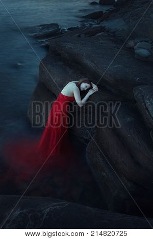A sad woman in red dress near the sea in evening. Long time exposure