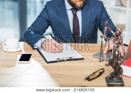 cropped shot of lawyer doing paperwork and using laptop at workspace in office
