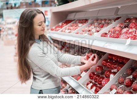 Cheerful happy customer girl choosing gifts for Christmas and New Year. She is taking red Christmas tree decoration balls. difficult decision. Various choice