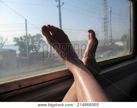 The female bare feet of the traveler on the train - conceptual photo. Women's bare legs lie on the window of an electric train and outside the window a village on the seashore