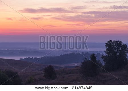Morning autumn landscape: fog over hills and forest on background of red sunrise. View from above. Village Opishnya Poltava region Ukraine