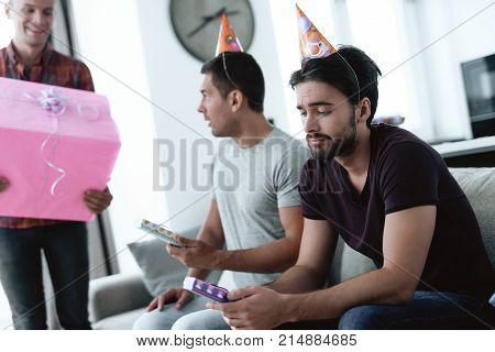 Men in birthday hats are showing to each other gifts. They are preparing for a birthday party. Two men are sitting on the couch with gifts in their hands.