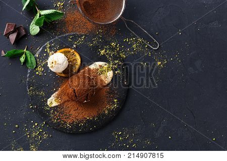Exquisite french dessert. Chocolate fondant with creme anglaise and vanilla ice cream on round slate with pistachio crumble. Black background with cocoa, mint and sieve, top view, copy space