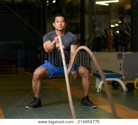 Strong Vietnamese athlete exerising in gym with battle ropes