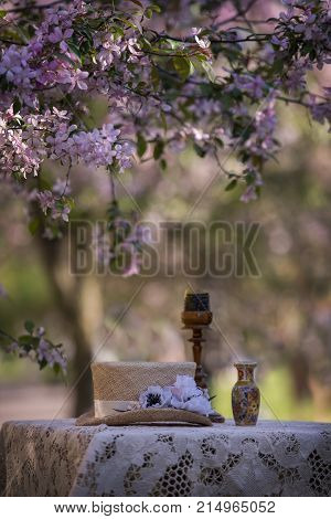 Still life in nature on the branches of a beautiful tree. The table is covered with a lace napkin on which the hat is a vase and a candlestick with a candle.