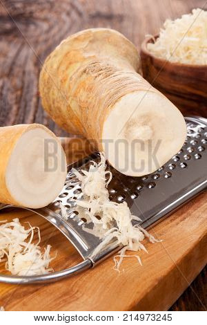 Fresh and grated horse-radish roots in wooden bowl on wooden background.