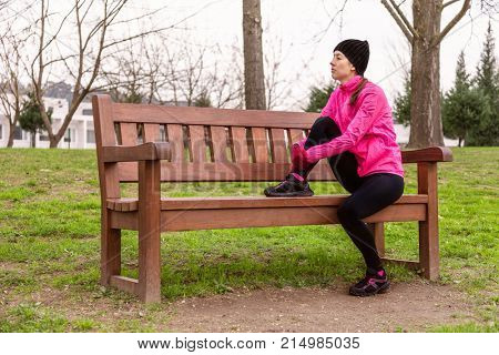 Female athlete tired or depressed resting on a bench on a cold winter day on the training track of an urban park. Young woman wearing pink windbreaker, beanie, gloves and running tights
