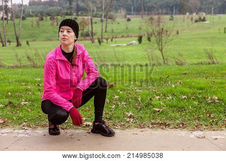 Young woman analyzing the track before running on a cold winter day on the training track of an urban park. Female athlete wearing pink windbreaker, beanie, gloves and running tights