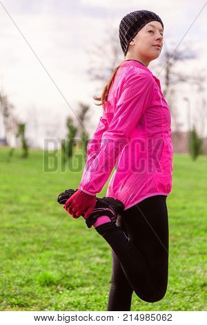Young woman warming up and stretching the legs before running on a cold winter, autumn of fall day in an urban park. Female athlete wearing pink windbreaker, beanie, gloves and running tights