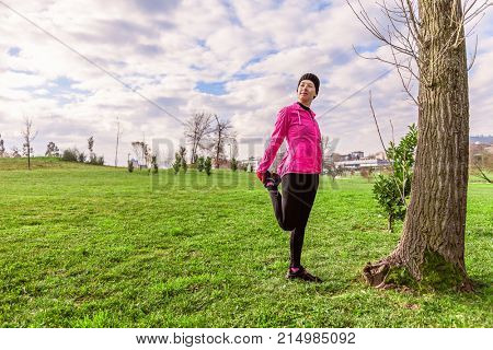 Young woman warming up and stretching the legs before running on a cold winter, autumn of fall day in an urban park. Female athlete wearing pink windbreaker, beanie, gloves and running tights