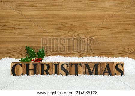 The word CHRISTMAS made from wooden block letters on some snow with a sprig of holly and a rustic wooden background with copy space to add your own message.