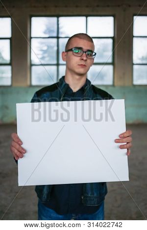 Young Man Holding White Background Frame. Teenager With A Confused Face, Out Of Focus.