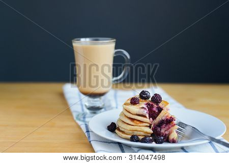 Eating Of Fritter Or Oladyi With Black Raspberry With Fork On White Plate And Cappuccino On Table Wi