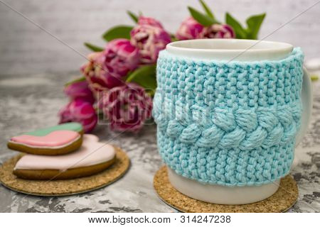 White Cup Of Tea In A Knitted Blue Case With Pink Flowers Tulips And Sweets, With Gingerbread On A T