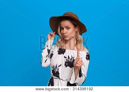 Grumpy Young Model With Curly Blond Hair In A Sundown Hat And White Dress Wags Finger Standing On A 