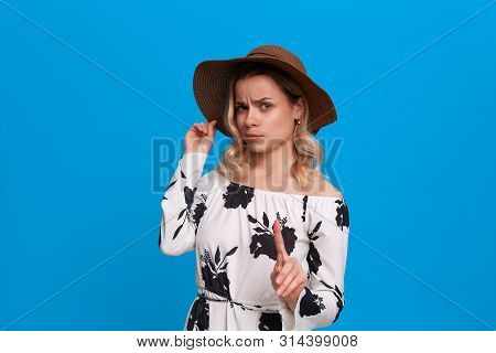 Grumpy Young Model With Curly Blond Hair In A Sundown Hat And White Dress Wags Finger Standing On A 