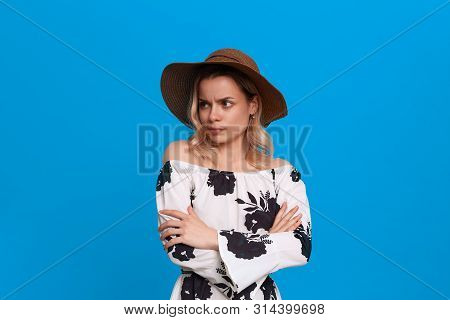 Grumpy Young Model With Curly Blond Hair In A Sundown Hat And White Dress Looks At The Camera Standi