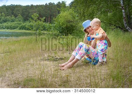 Happy Mom And Her Little Baby Are Playing And Communication By The Lake In A Summer Green Forest