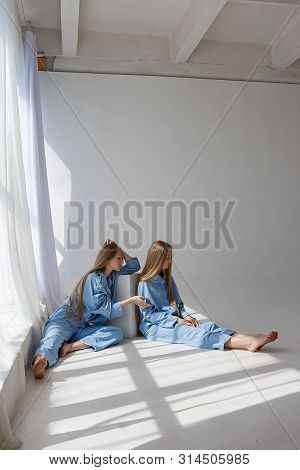 Two Young Pretty Twin Girls In Identical Blue Suits With Long Blond Hair Sitting On White Cyclorama 
