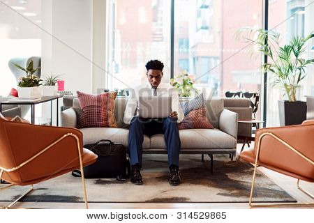 Businessman Sitting On Sofa Working On Laptop At Desk In Shared Workspace Office