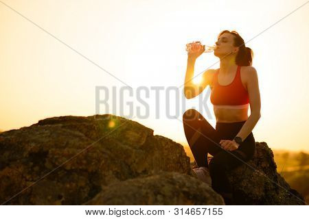 Young Woman Resting and Drinking Water after Trail Running Outdoor. Workout Exercise at Hot Beautiful Summer Sunset. Sport and Healthy Active Lifesyle Concept.