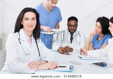 Female Doctor At Concilium, Looking At Camera. Medical Team On Background