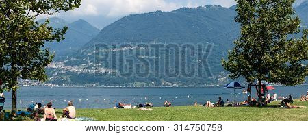 Lake Como, Italy - July 21, 2019: People Resting On Beach Near Lake. Trees And Alp Mountains On Back