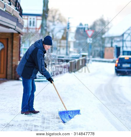 Man With Snow Shovel Cleans Sidewalks In Winter During Snowfall. Winter Time In Europe. Young Man In