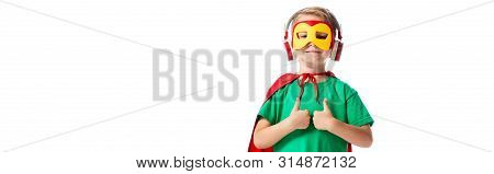 Panoramic Shot Of Smiling Boy In Hero Costume Listening Music In Headphones And Showing Thumbs Up Is
