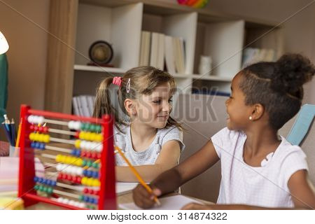 Two Little Girls Sitting At A Desk, Writing In Their Notebooks, Doing A Math Homework And Studying F