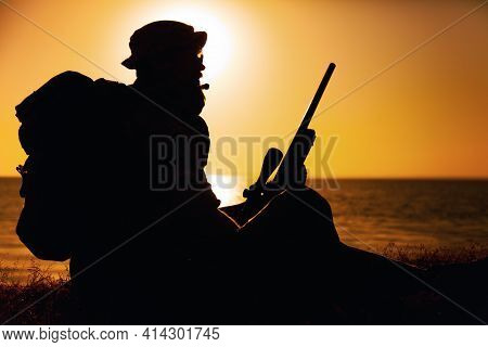 Silhouette Of Commando Fighter, Army Special Forces Sniper Sitting On Sea Or Ocean Shore On Sunset. 