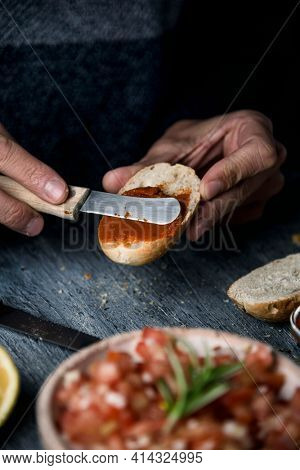 a young caucasian man spreads a vegan version of the spanish sobrasada, made with sun-dried tomato and almonds, on a toasted halved bread bun, on a gray table with a tomato salad on the foreground