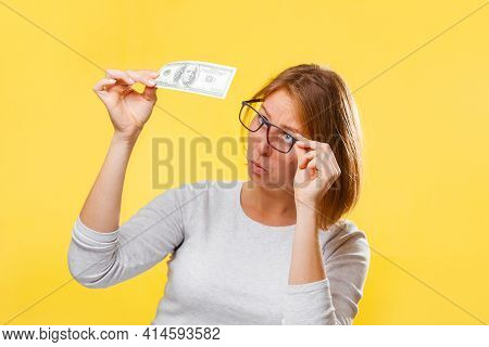Portrait Of Woman Looking At The Dollar Bill Checking For Falsity. Yellow Background. The Concept Of