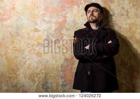 attractive young bearded man in a pea coat and cap is standing at the wall