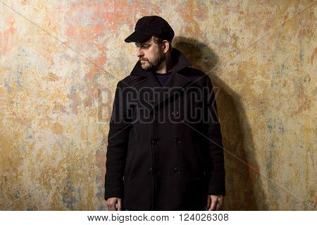 attractive young bearded man in a pea coat and cap is standing at the wall