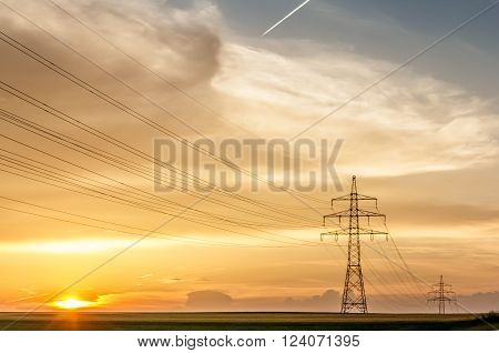 transmission line with poles in front of a yellow and amber sky at sunset or sunrise with condensation trail