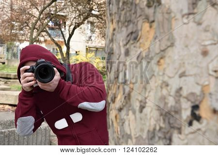 Mysterious paparazzi photographer with the hood on his head lurking behind the tree