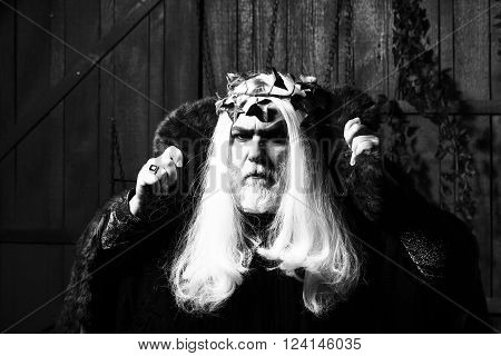 Bearded senior man in long white wig vine crown as Zeus god in fur coat indoor on wooden background