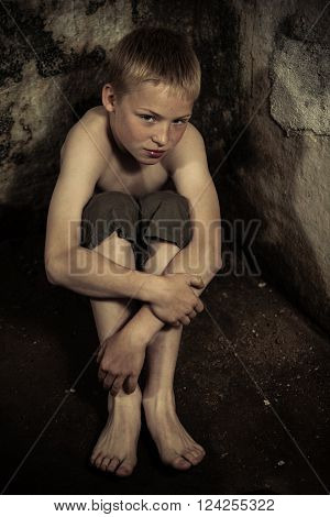 Sad single imprisoned male child wearing shorts sitting on dirty floor in dark, damp dungeon with stone walls