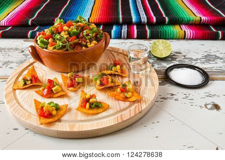 Nachos chips and vegetables in an earthenware bowl and tequila with a poncho on background over a wooden table