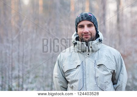 Portrait of a handsome man with rugged stubble, wearing warm clothes while standing outdoors on a cold winter day, looking at the camera with a subtle smile and a snowy forest behind him
