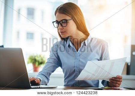 Double-checking everything. Young beautiful woman in glasses using laptop and holding documents while sitting at her working place