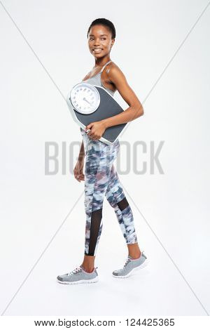 Full legth portrait of a happy fitness afro american woman holding weighing machine isolated on a white background