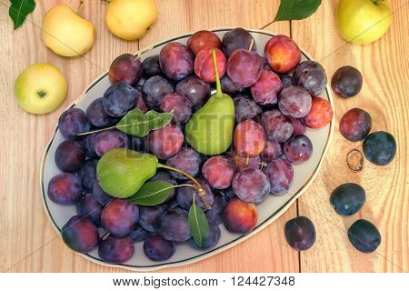 On the surface of the wooden table is a ceramic dish filled with large Mature plums pears. Next to them are apples.