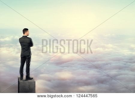 Young man in business suit, standing on a concrete column above the clouds. Business, leadership and success concept