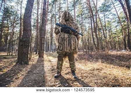 Sniper in camouflaged ghillie suit standing in forest with rifle in hands.Low angle view.Selective focus.