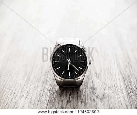 Silver reflective wrist watch clock with black dial and silver pointers resting on wood surface. Selective focus on the clock.
