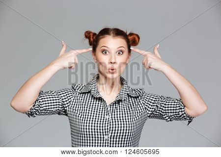 Joyful comical young woman pointing on her temples and making funny face over grey background