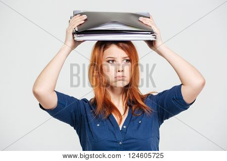 Tired sad redhead young woman standing with folders on her head over white background