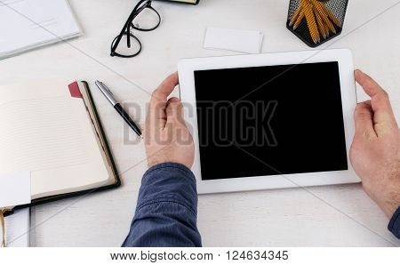 Man holding tablet with blank screen while sitting at the white wooden table. Man communicates using a tablet computer